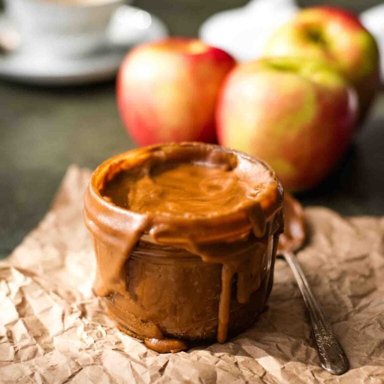 A square shot of bourbon butterscotch sauce in a glass jar sitting on brown paper with a spoonful of sauce to the side. In the background there is a white napkin, cup of coffee, and fresh apples.