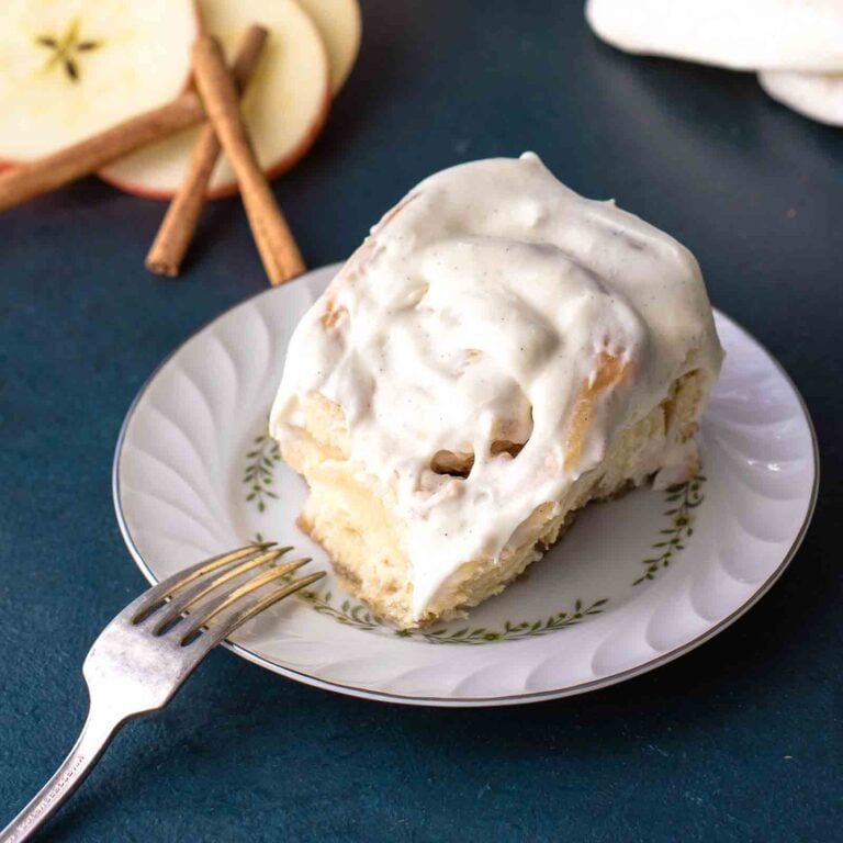 An apple cinnamon roll with vanilla bean cream cheese icing on a white plate with a blue background.