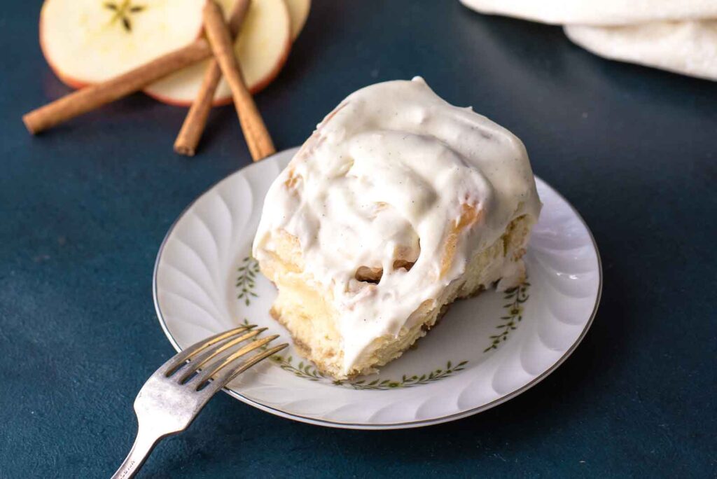 A single cinnamon roll with white frosting on a decorative plate is placed next to a fork. In the background, there are apple slices and cinnamon sticks, making it a perfect treat for kids.