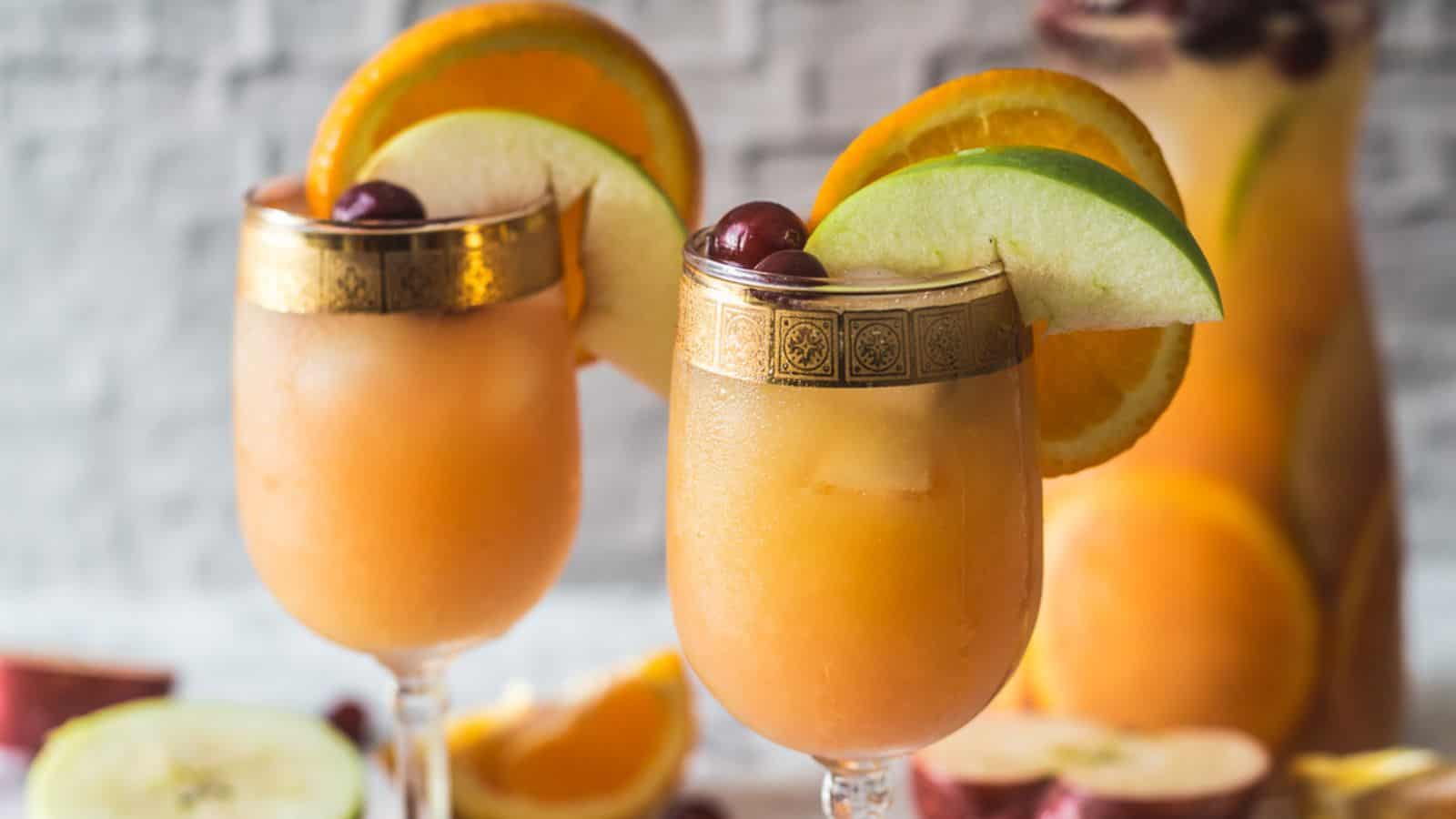 Two glasses of orange-colored cocktails garnished with apple slices, orange slices, and cherries. A pitcher filled with similar incredible drinks is in the background, along with assorted fruit.