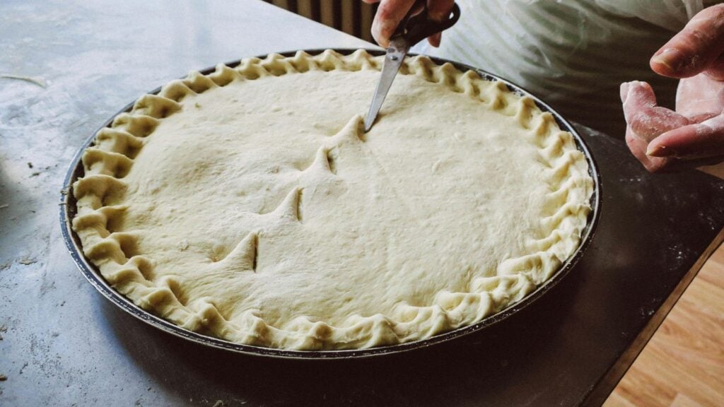 A person using scissors to cut vents into a pie crust with fluted edges. The pie is set on a countertop, with a second hand poised to adjust the dough.