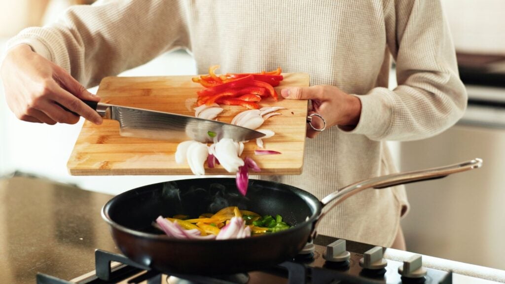 A person using a knife to push ingredients from a wooden chopping board to a frying pan.