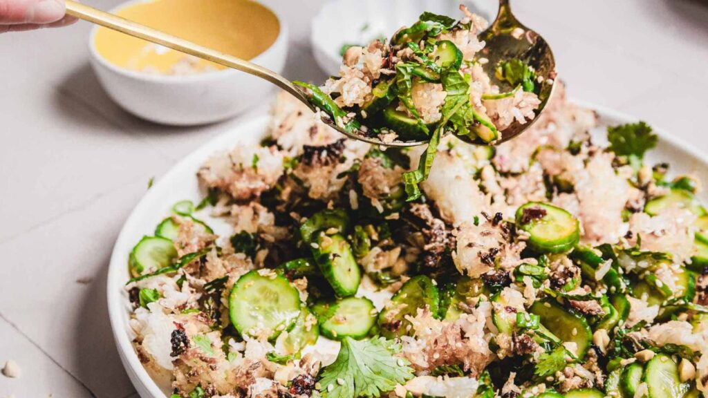 A close-up of a dish featuring rice, sliced cucumbers, herbs, and pieces of a pink ingredient, possibly grapefruit. A spoon is lifting a portion from the plate. The background shows a small bowl with a yellow interior.
