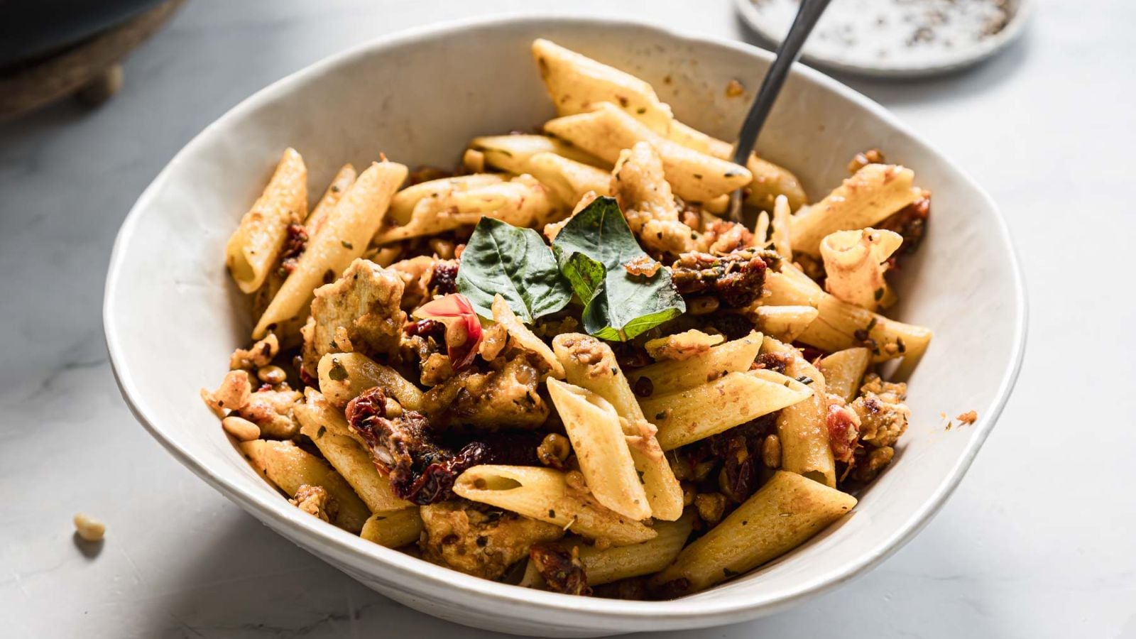 A white bowl filled with penne pasta mixed with chunks of chicken, sun-dried tomatoes, and garnished with fresh basil leaves. A fork is placed in the bowl, and a small dish is visible in the background.