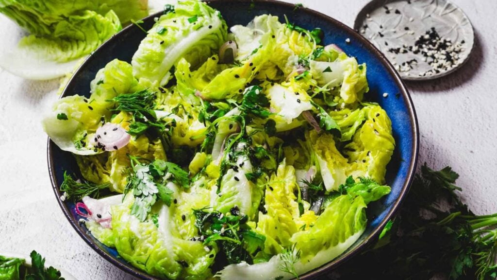 Un bol bleu rempli d'une salade verte contenant de la laitue frisée, des herbes fraîches et des oignons rouges tranchés. La salade est garnie de graines de sésame noires.