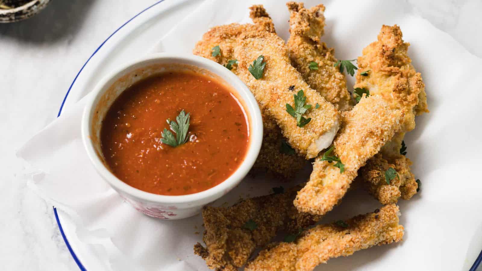 A plate of breaded chicken tenders is arranged around a small bowl of red dipping sauce, garnished with a sprig of parsley, on a paper-lined plate.