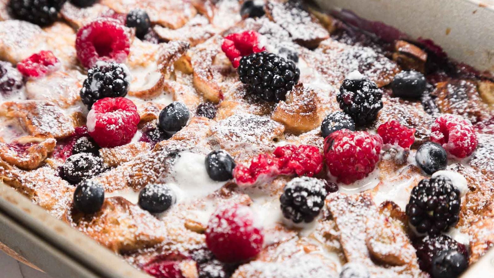 Close-up of a baked dessert topped with powdered sugar, featuring blackberries, raspberries, and blueberries. The dish is in a rectangular baking pan, with visible golden-brown edges and drizzles of cream or sauce.