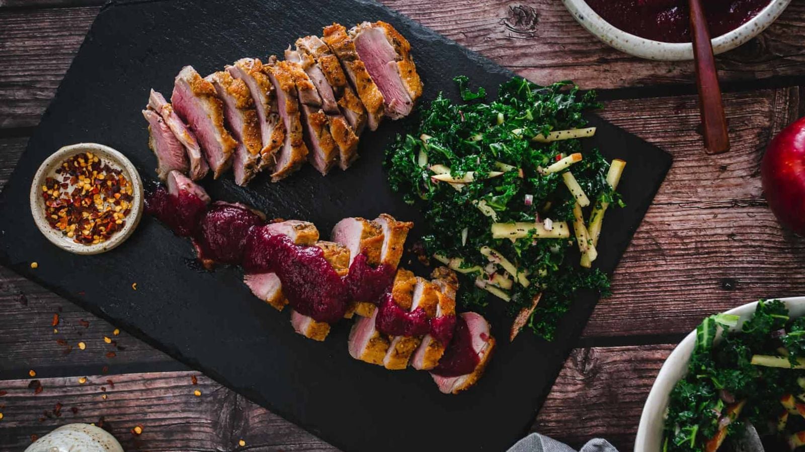 Sliced duck breast with a dark sauce is served on a black slate. Accompanied by a side of kale salad and a small bowl of red pepper flakes. The wooden table background adds a rustic touch.