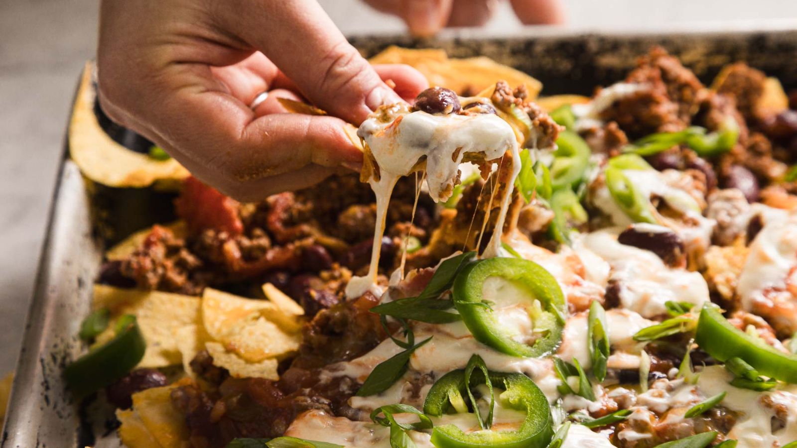 A close-up of nachos topped with melted cheese, ground beef, sliced jalape&ntilde;os, chopped green onions, sour cream, and beans. The tortilla chips are spread out with the toppings generously applied, creating a colorful and appetizing presentation.