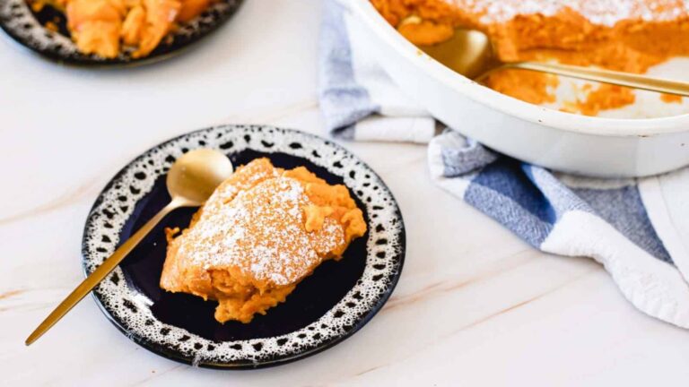 A serving of pumpkin souffle on a black plate with a gold spoon, dusted with powdered sugar. More souffle in a baking dish is visible in the background.