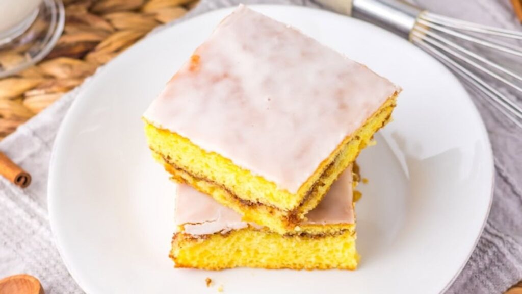Two slices of frosted yellow cake are stacked on a white plate, placed on a textured tablecloth. A whisk and wooden utensils are visible in the background.