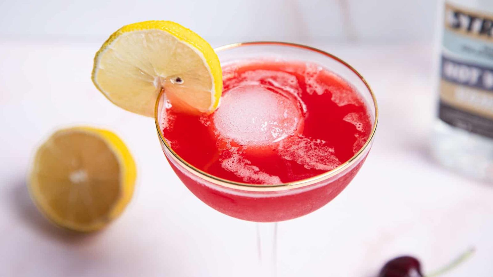 A mocktail in a glass with a gold rim is filled with a red liquid and a large round ice cube. A lemon slice is on the rim. A halved lemon and a cherry are nearby on a white surface.