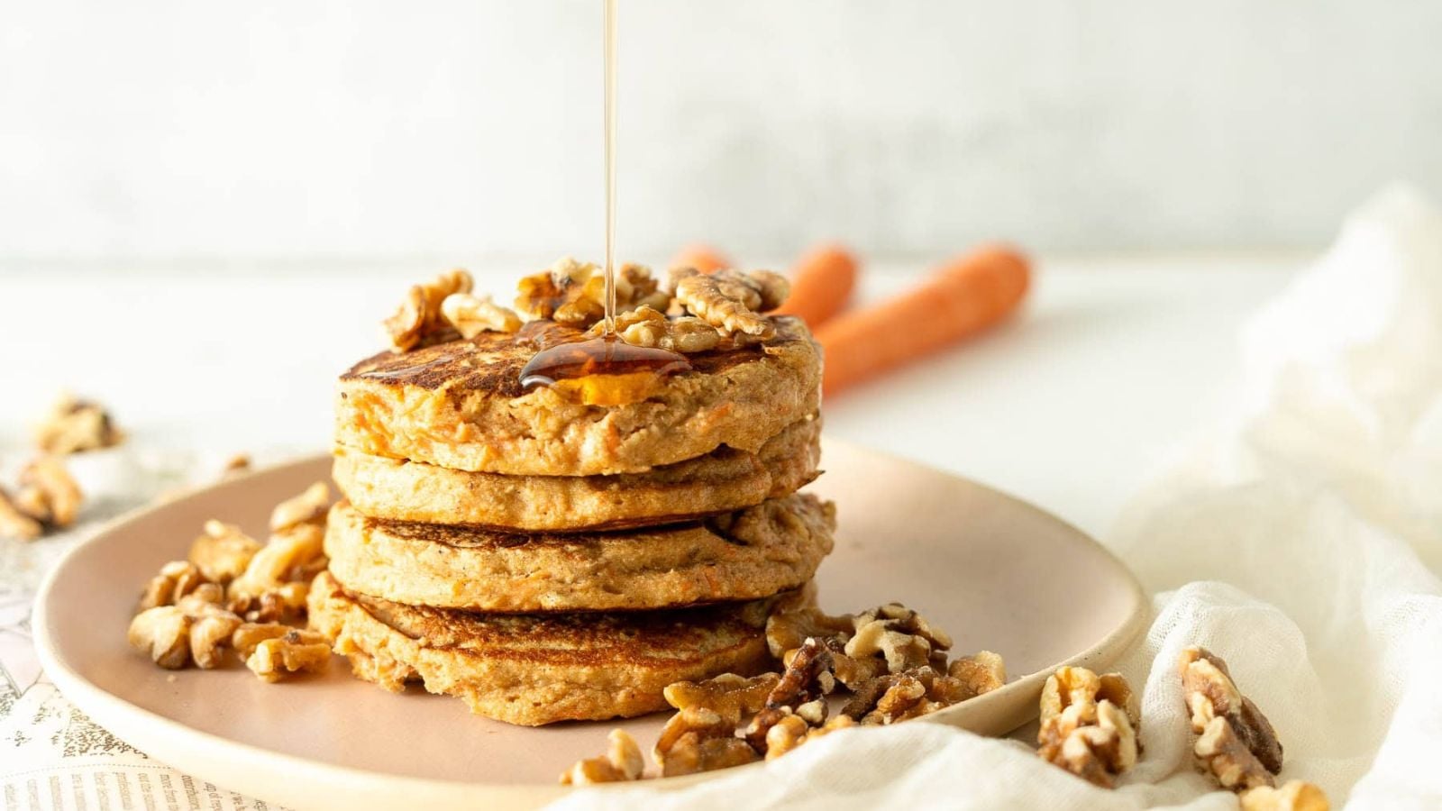 A stack of three pancakes topped with walnuts is on a pink plate. Syrup is being drizzled over the pancakes. Additional walnuts are scattered around the plate. Two carrots are partially visible in the background.