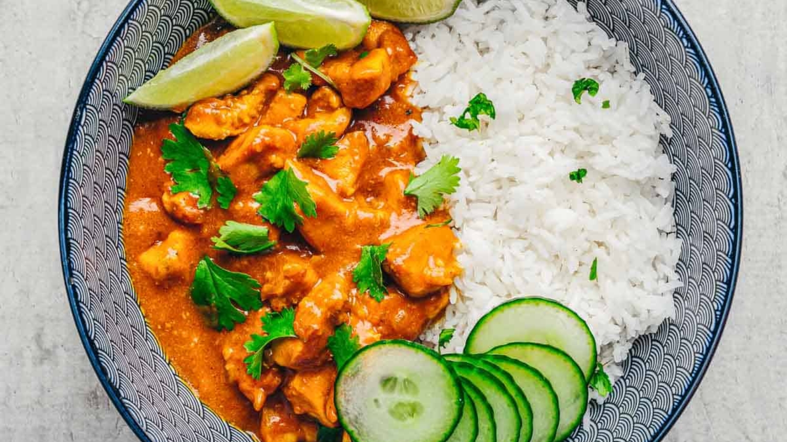 Bowl of curry with chicken, white rice, lime wedges, cucumber slices, and fresh cilantro on top.