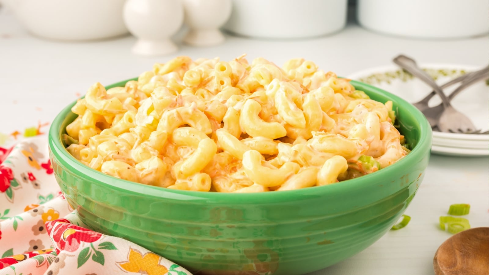 A green bowl filled with creamy macaroni and cheese sits on a table next to a floral napkin, a fork, and a spoon&mdash;an irresistible choice for Memorial Day BBQ dishes.
