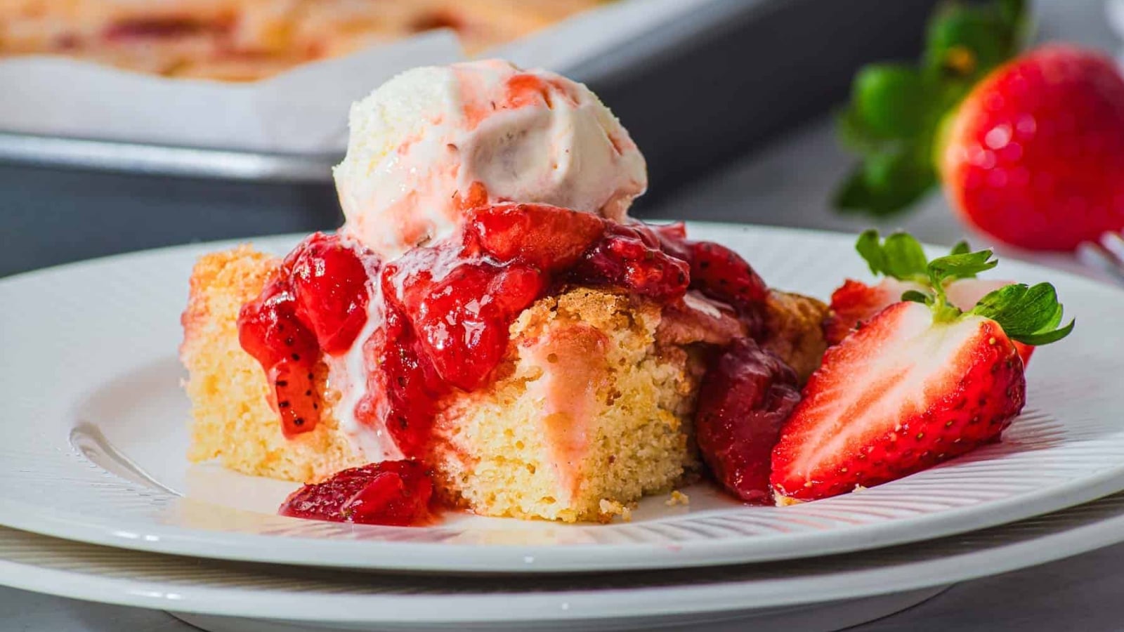 A slice of cake topped with strawberry sauce and a scoop of vanilla ice cream sits on a white plate, with a halved fresh strawberry beside it. A baking tray and whole strawberries are in the background.