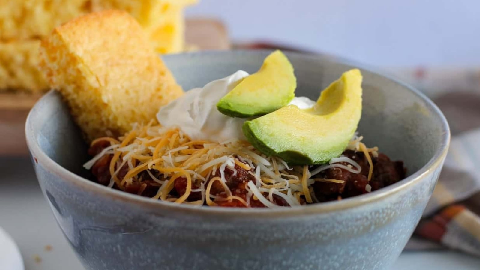 A bowl of chili topped with shredded cheese, sour cream, and avocado slices, accompanied by pieces of cornbread.
