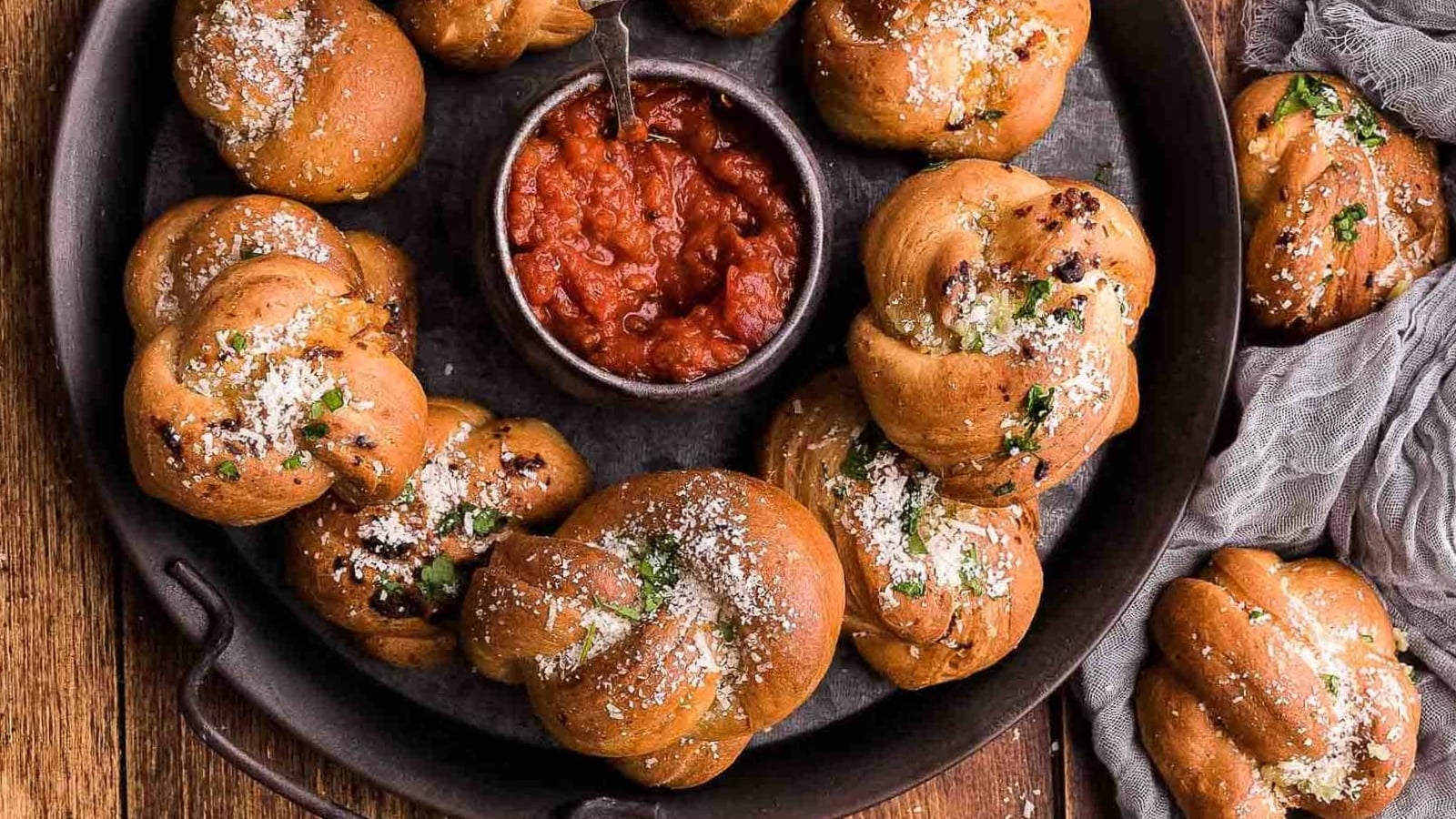 A round tray holds several baked garlic knots sprinkled with herbs and cheese, arranged around a bowl of marinara sauce for dipping.
