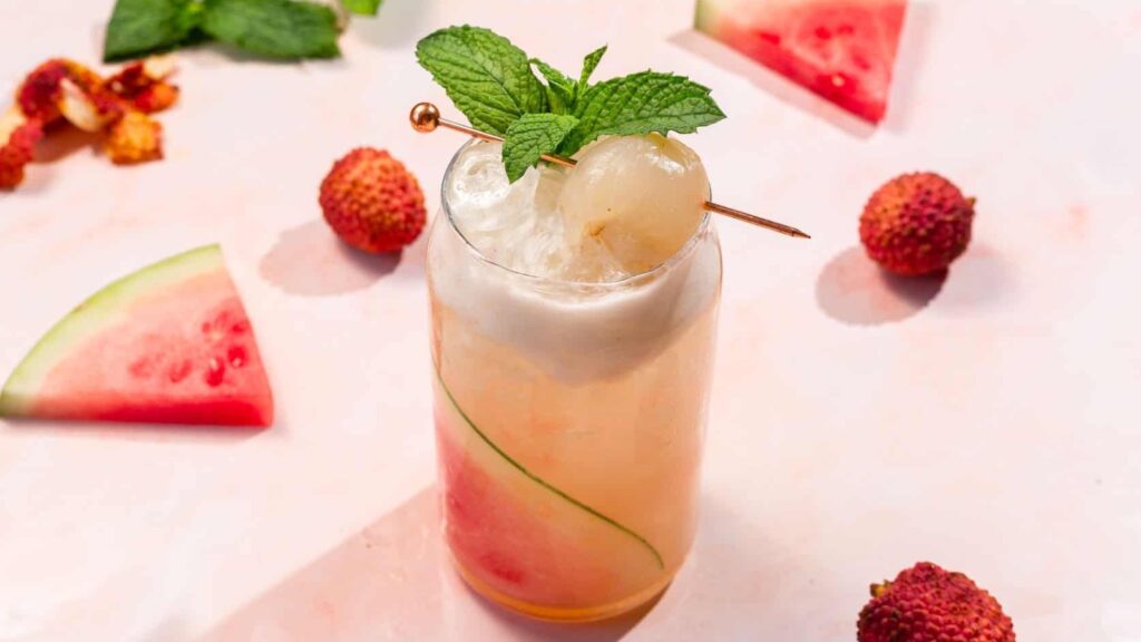A tall glass of lychee and watermelon cocktail with ice, garnished with mint and lychee, surrounded by watermelon slices and lychees on a light surface.