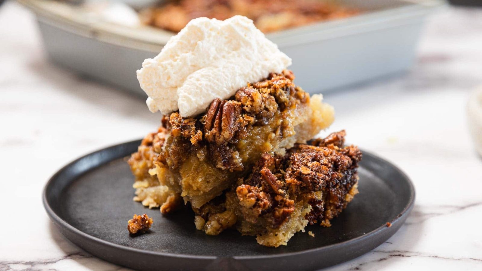 A slice of pecan pie topped with whipped cream on a black plate, with a baking dish in the background.