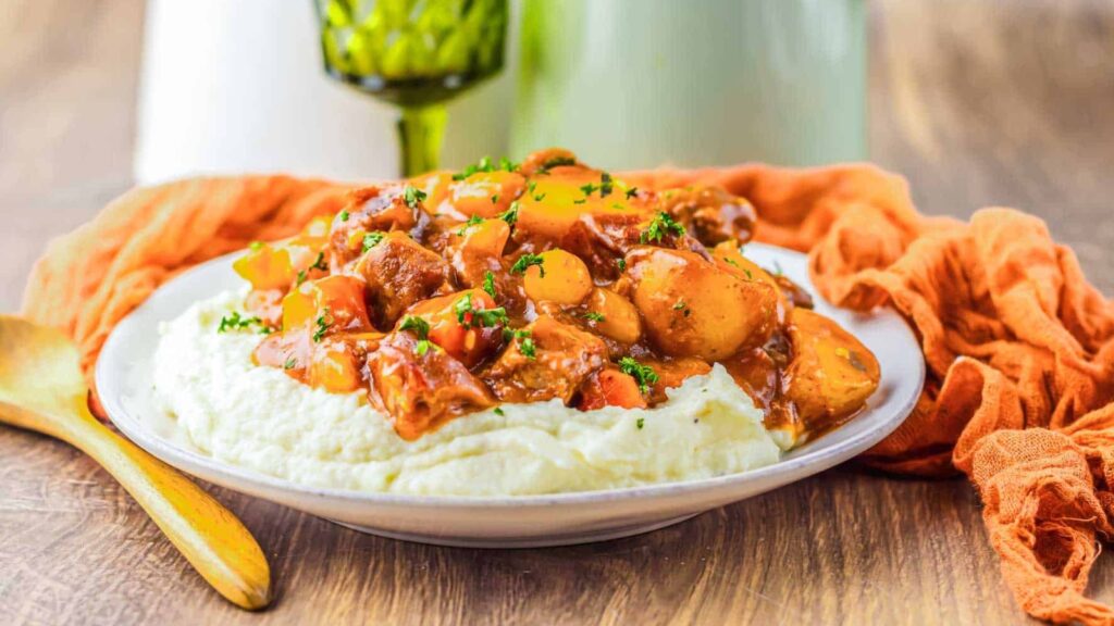 A plate of mashed potatoes topped with beef stew and vegetables, garnished with chopped parsley, sits on a wooden table beside a wooden spoon and an orange napkin.