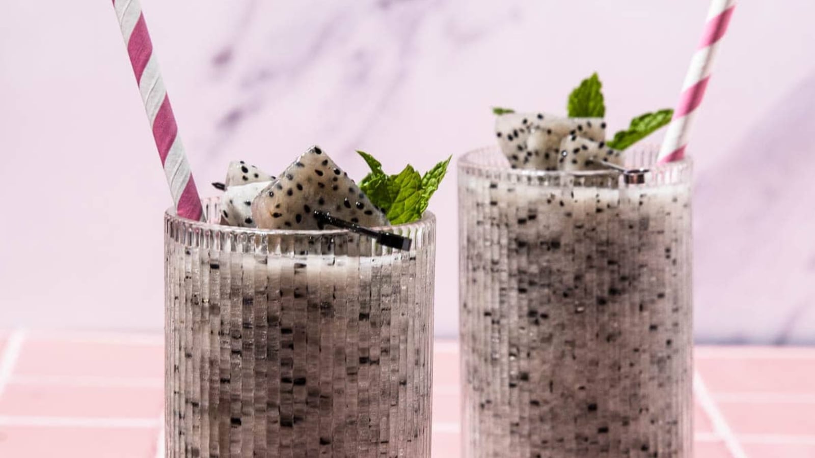 Two glasses of dragon fruit smoothie with mint garnish and pink striped straws on a pink tiled surface.