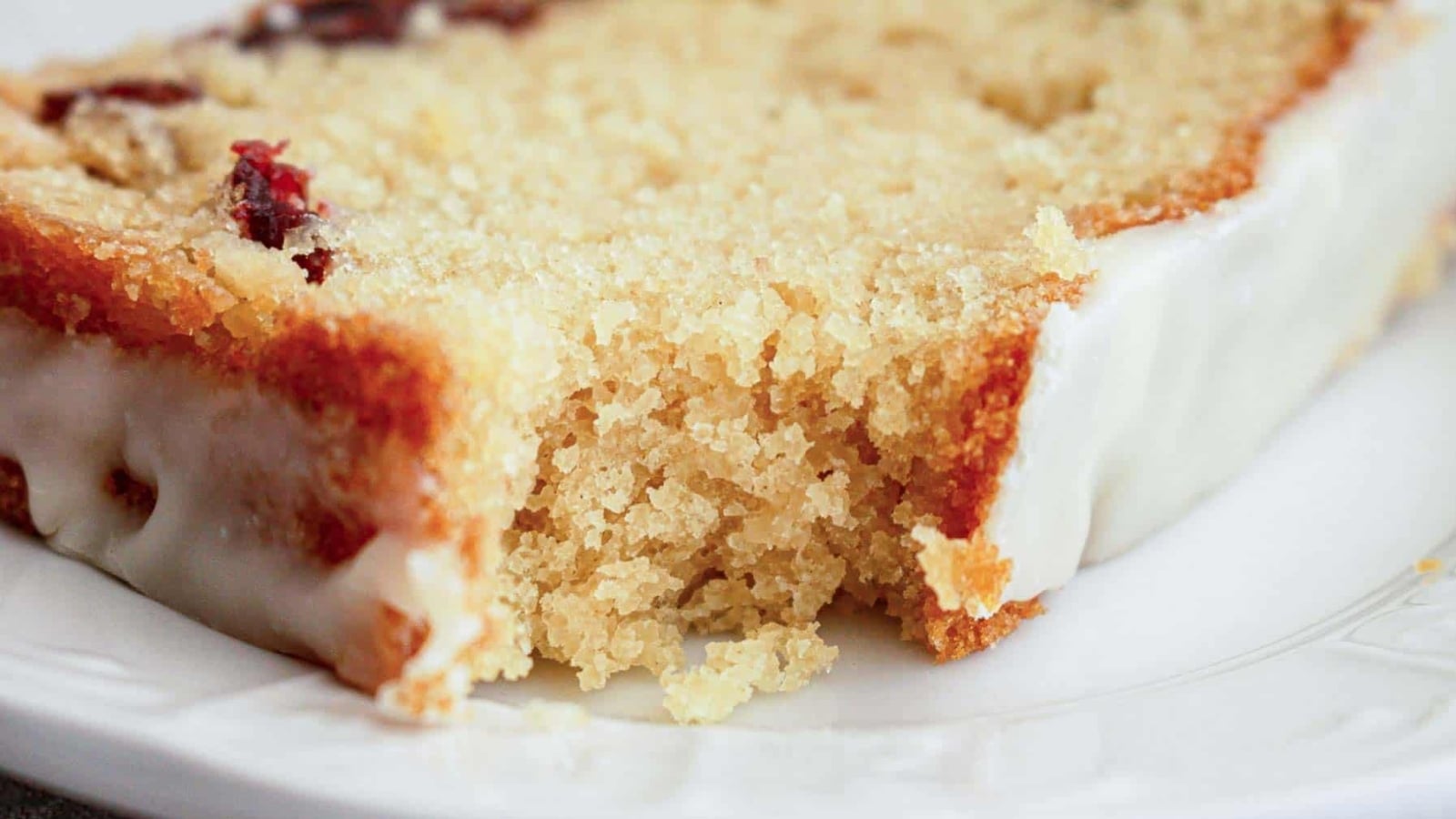 A loaf cake with white icing drips on a wooden board, next to two forks and some dried fruit pieces.