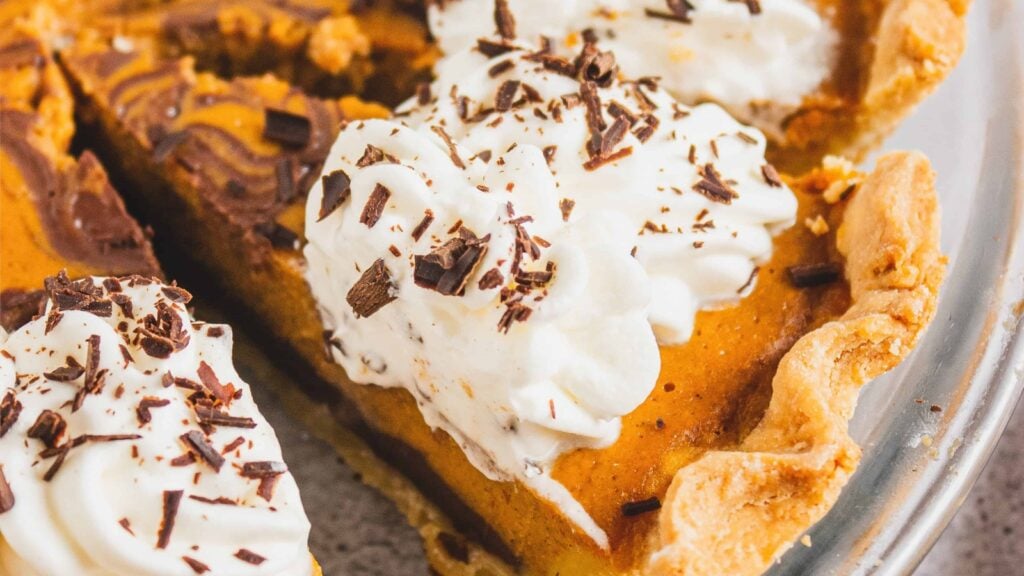Close-up of pumpkin pie slices topped with whipped cream and chocolate shavings in a metal pie dish.