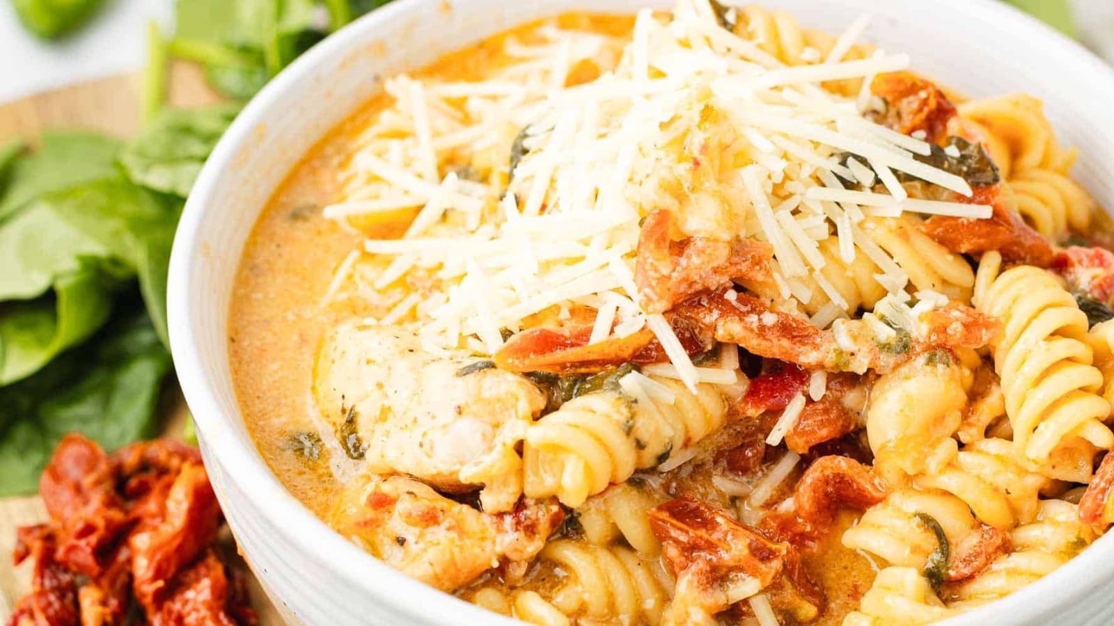 A bowl of creamy pasta with chicken, sun-dried tomatoes, spinach, and shredded cheese on top.