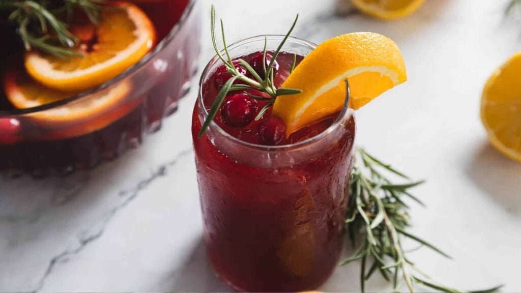 A glass of red punch with ice, garnished with an orange slice, cranberries, and a sprig of rosemary on a marble surface.