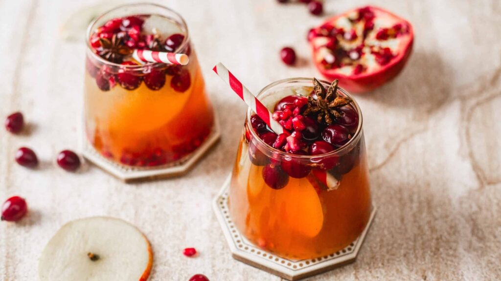 Two glasses of fruit-infused drink with red berries, pomegranate seeds, sliced fruit, and striped straws, garnished with star anise, on a light surface with scattered fruit.