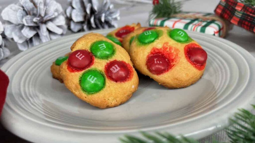 Two cookies topped with red and green M&M candies sit on a gray plate, with pine cones and a wrapped gift in the background.
