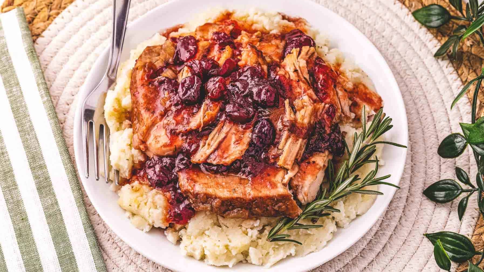 Shredded pork with cranberry sauce served over mashed potatoes, garnished with rosemary on a white plate.