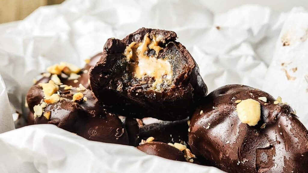 Close-up of chocolate-covered candies with a creamy filling, some topped with chopped nuts, placed on crumpled white parchment paper.