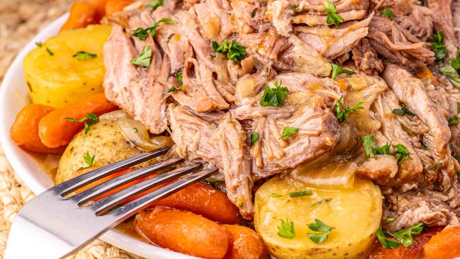 A bowl of shredded pot roast with baby potatoes and carrots, garnished with parsley, on a woven placemat.