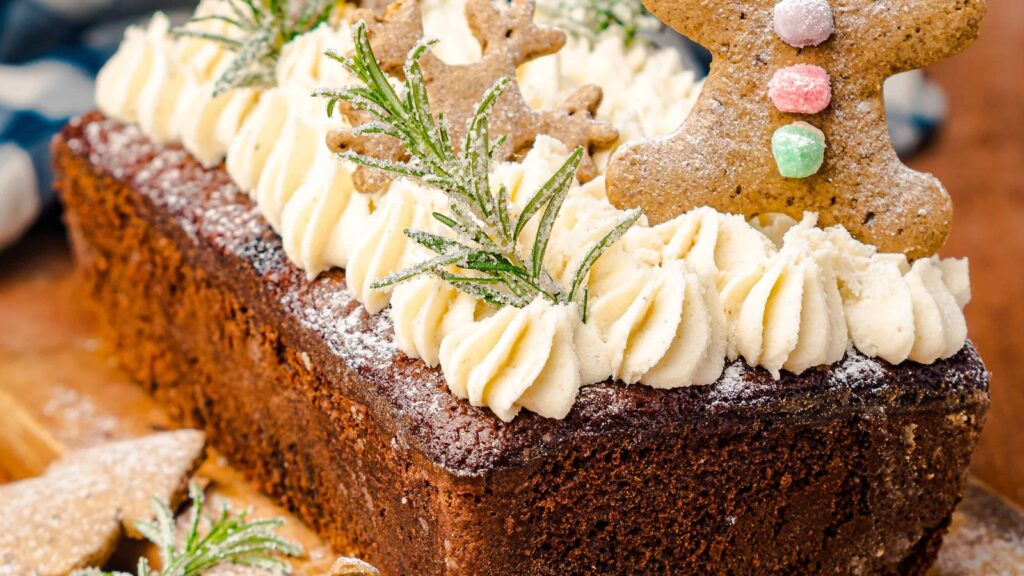 A rectangular chocolate cake with piped white frosting, rosemary sprigs, powdered sugar, and gingerbread decorations on top.