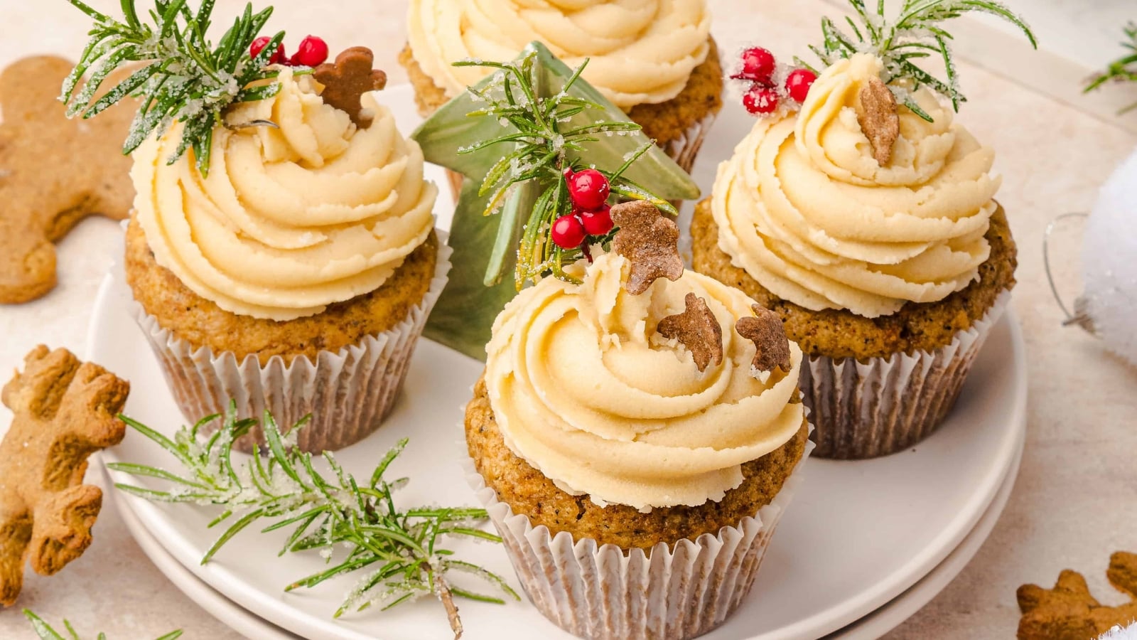 Cupcakes with cream frosting, rosemary, berries, and gingerbread cookies on white plates, festive background.