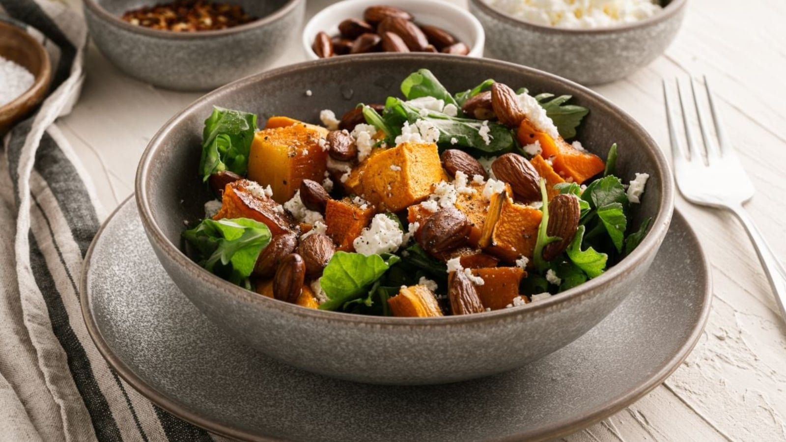 A bowl of salad with roasted pumpkin, leafy greens, almonds, feta cheese, and a bowl of almonds and other condiments in the background.