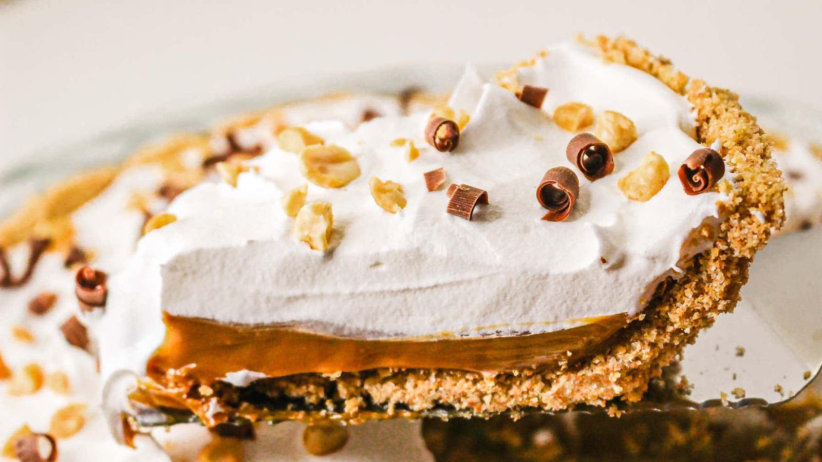 A close-up of a creamy pie slice with whipped topping, chocolate curls, and chopped nuts in a crumb crust.