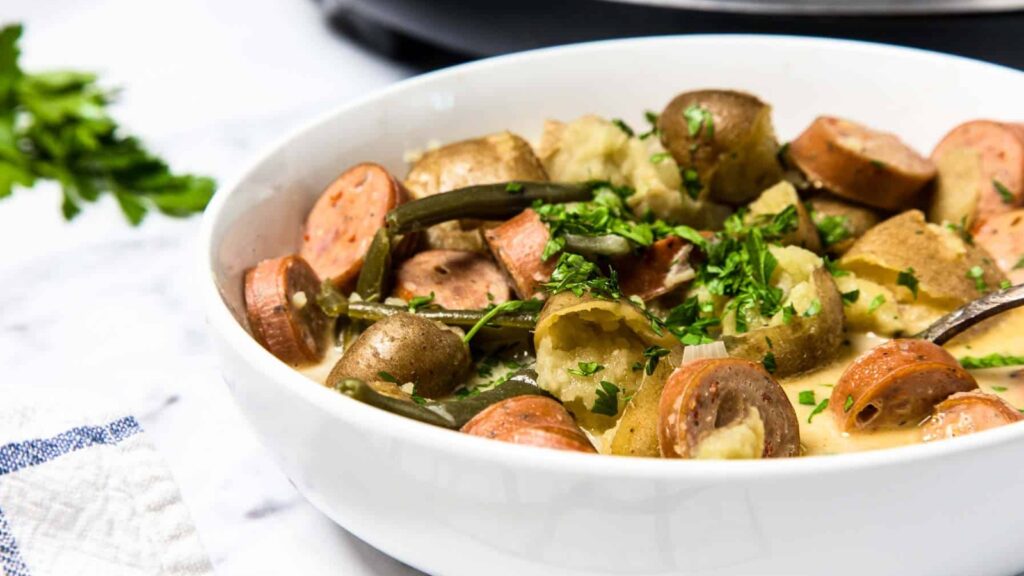 A white bowl filled with sliced sausage, potatoes, green beans, and chopped parsley in a creamy sauce, on a marble surface.