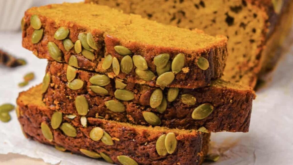 Three slices of pumpkin bread topped with pumpkin seeds are stacked on a piece of parchment paper, with the rest of the loaf in the background.