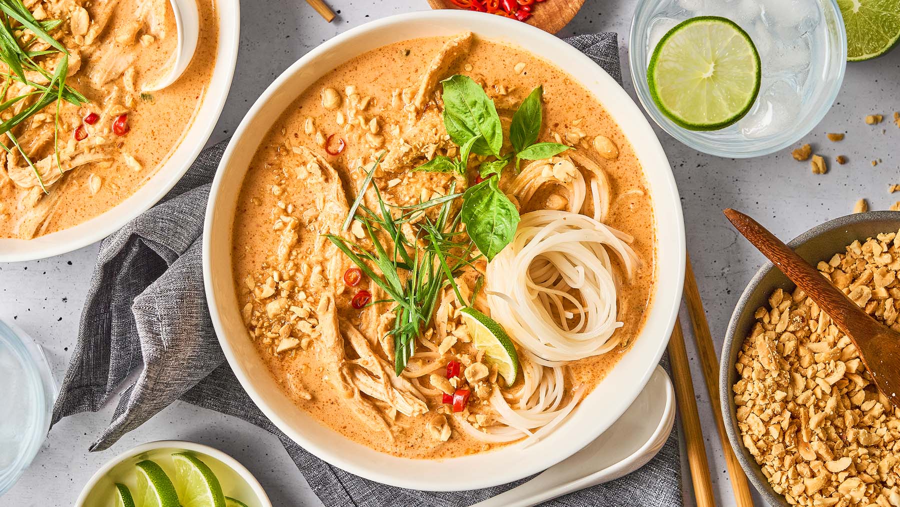 A bowl of noodle soup with shredded chicken, creamy broth, fresh herbs, chopped peanuts, sliced chili, and lime wedges, surrounded by additional garnishes and a glass of water.