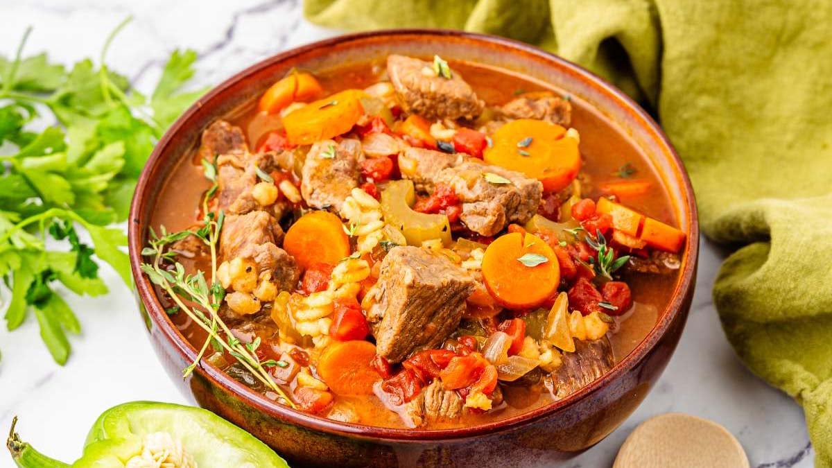 A bowl of beef and vegetable stew with chunks of meat, carrots, tomatoes, and herbs, placed on a table next to parsley, a green pepper, and a green cloth.