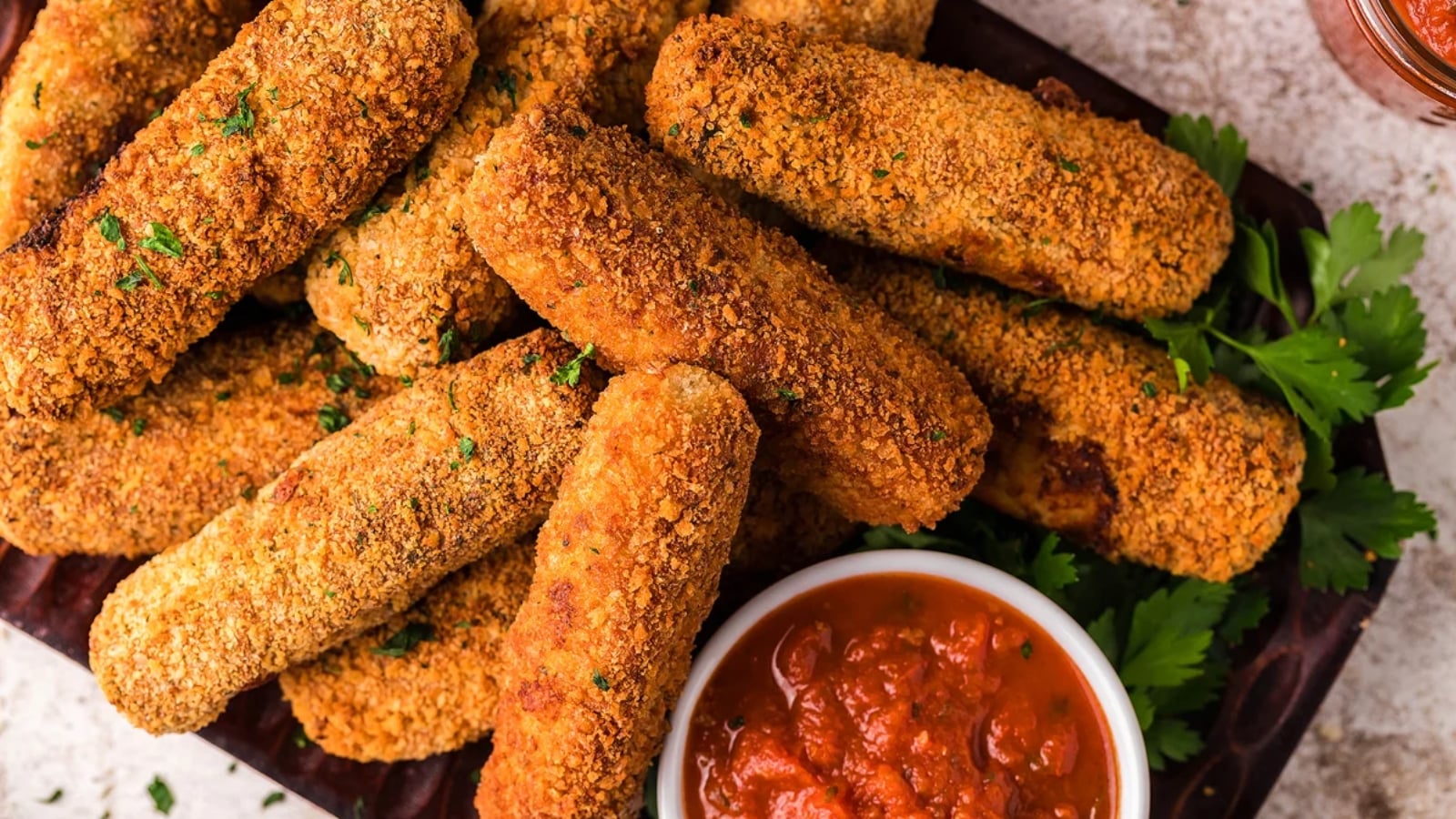 A plate of breaded mozzarella sticks is arranged around a bowl of marinara sauce, garnished with parsley.