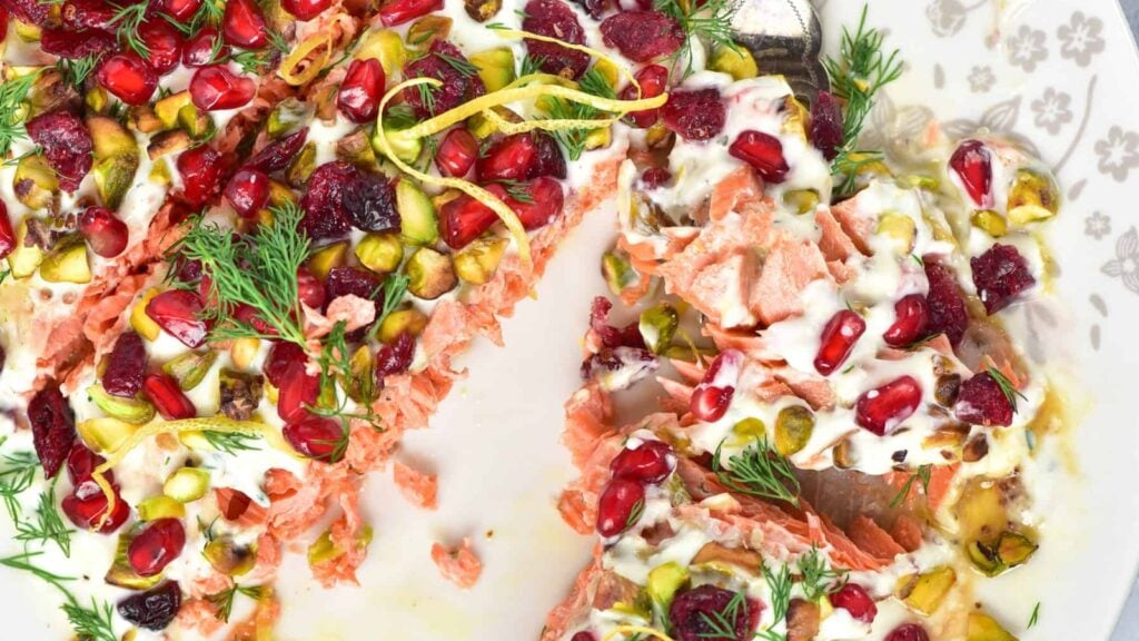 A baked salmon fillet topped with yogurt sauce, pomegranate seeds, pistachios, dried cranberries, lemon zest, and fresh dill on a white plate.