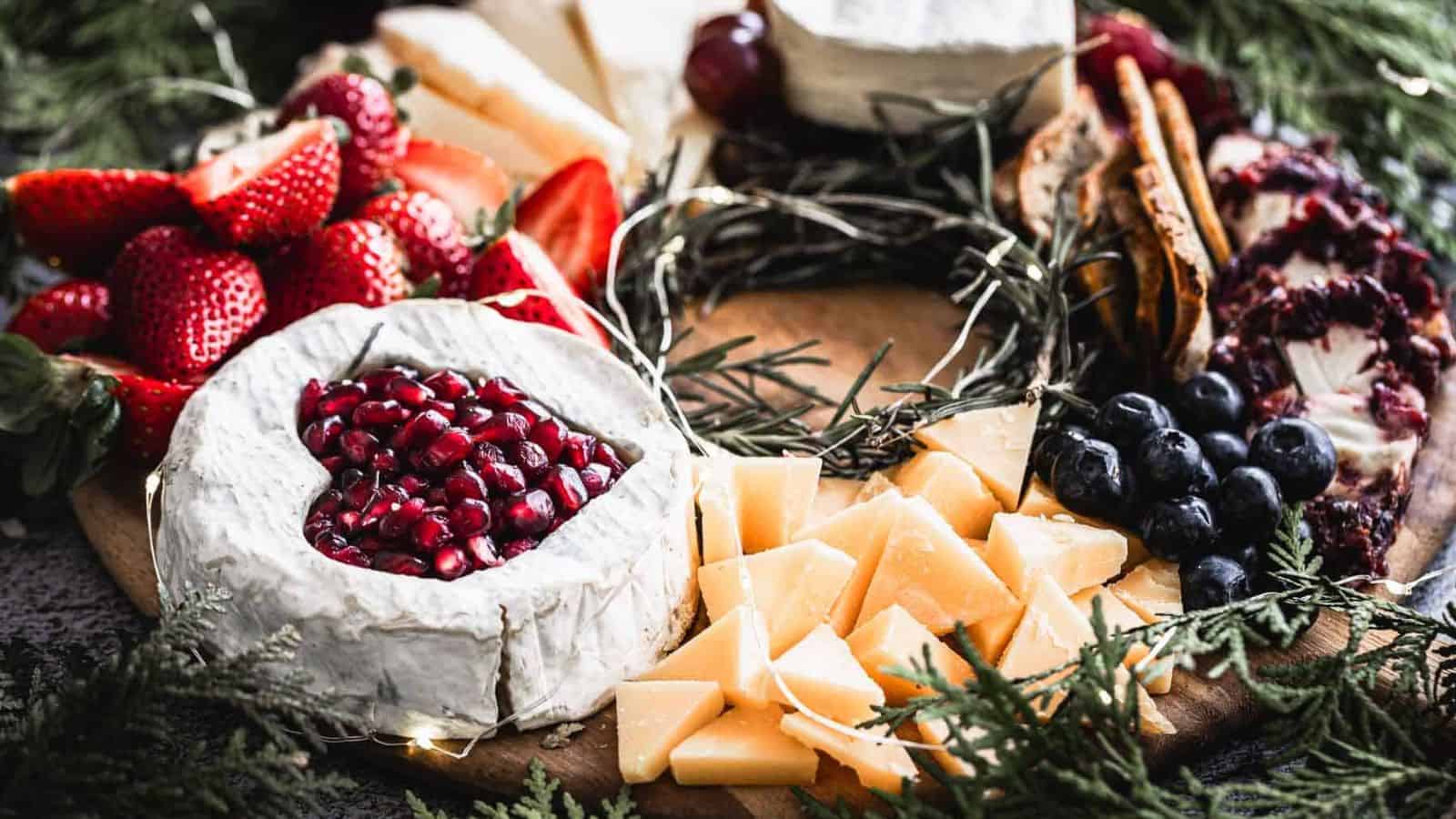 A charcuterie board with heart-shaped cheese topped with pomegranate seeds, surrounded by sliced strawberries, blueberries, grapes, assorted cheeses, crackers, and pine boughs.
