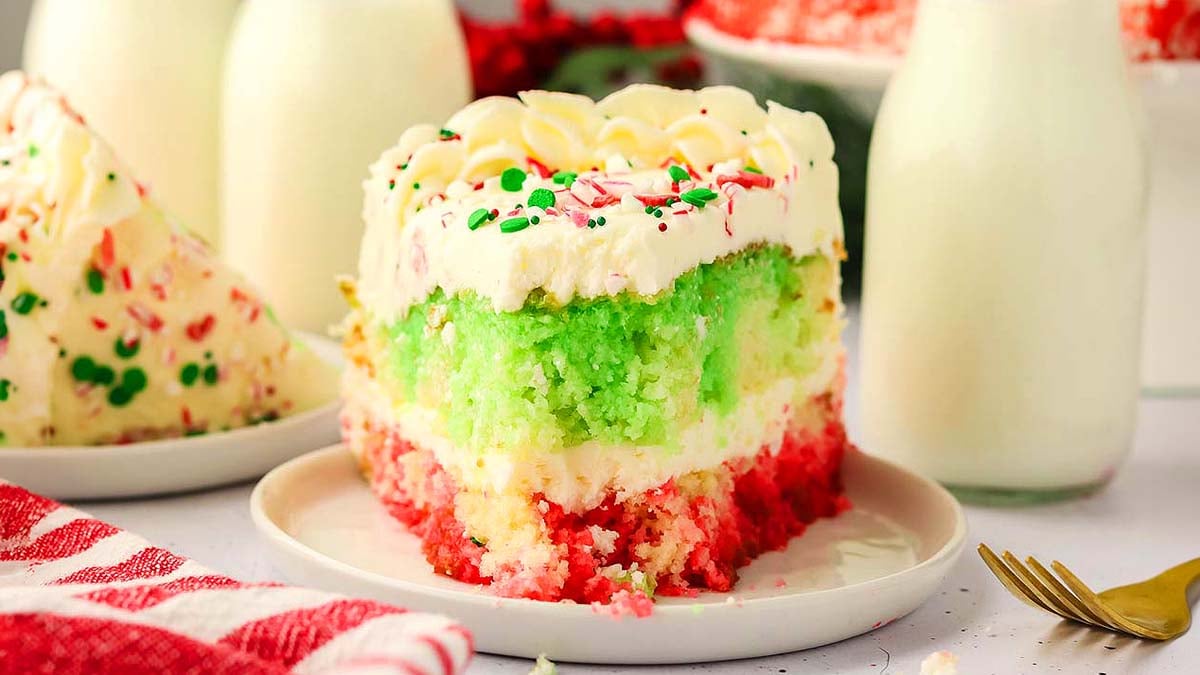 A slice of layered red, green, and white cake with white frosting and sprinkles on a white plate next to a bottle of milk.
