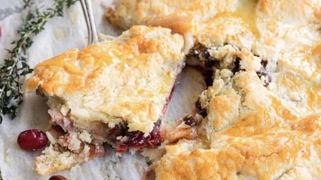 A close-up of a sliced fruit pie with a golden, flaky crust, exposing a filling of mixed berries or cherries. A serving utensil is partially visible.