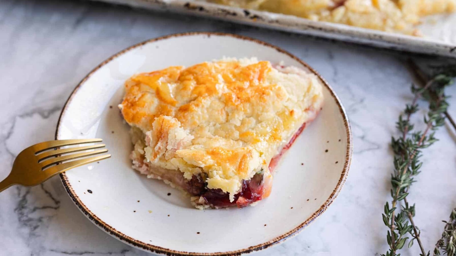 A slice of fruit pie with a golden crust on a white plate, next to a gold fork and fresh thyme.