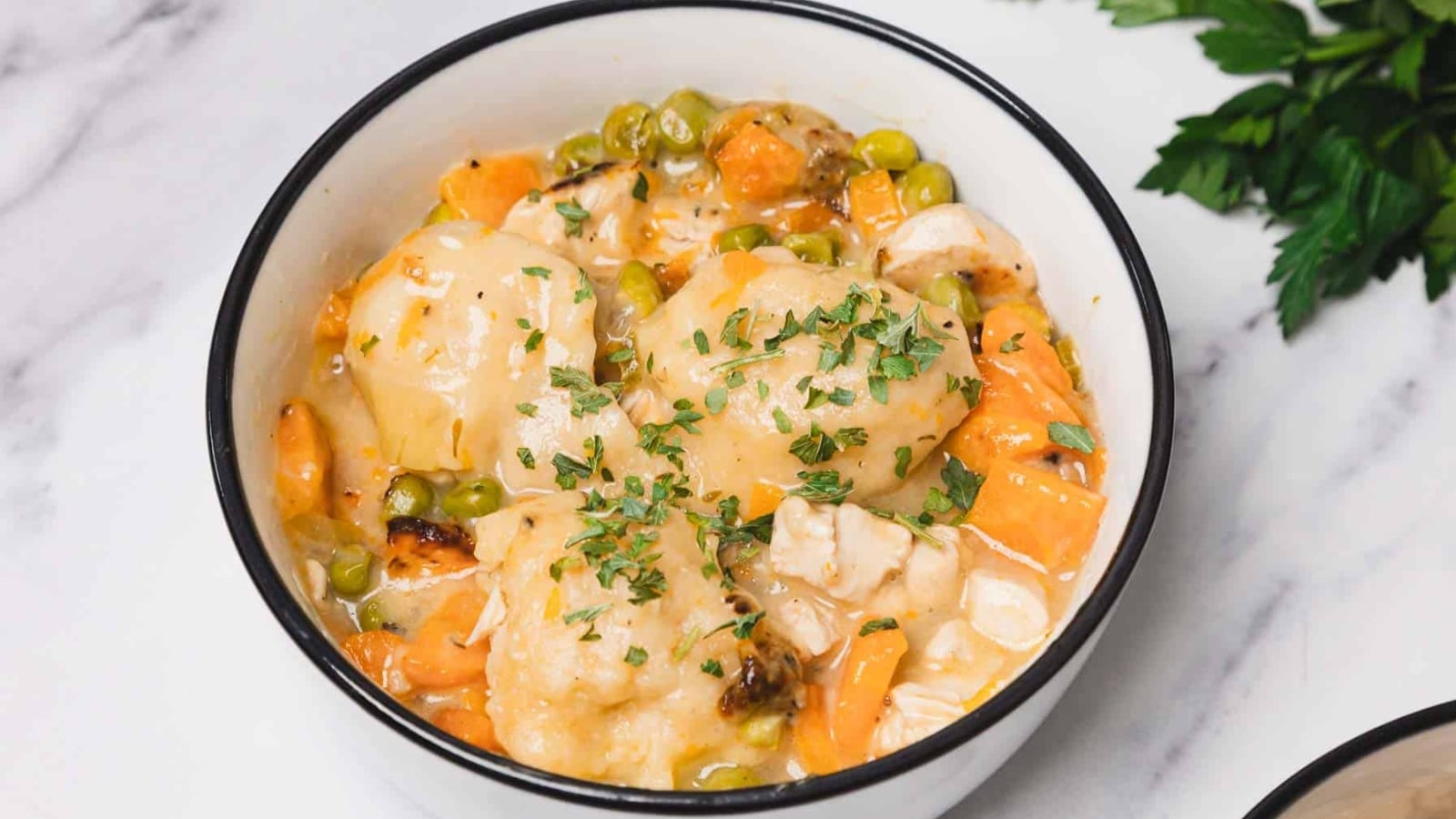 A bowl of chicken and dumplings soup with carrots, celery, and herbs, with a spoon inside.